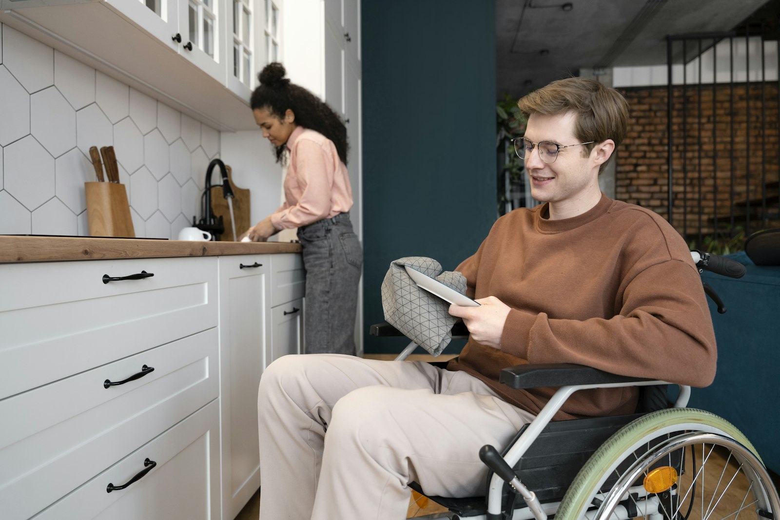 A wheelchair user reading a tablet at his kitchen table while another person prepares food at the counter behind him